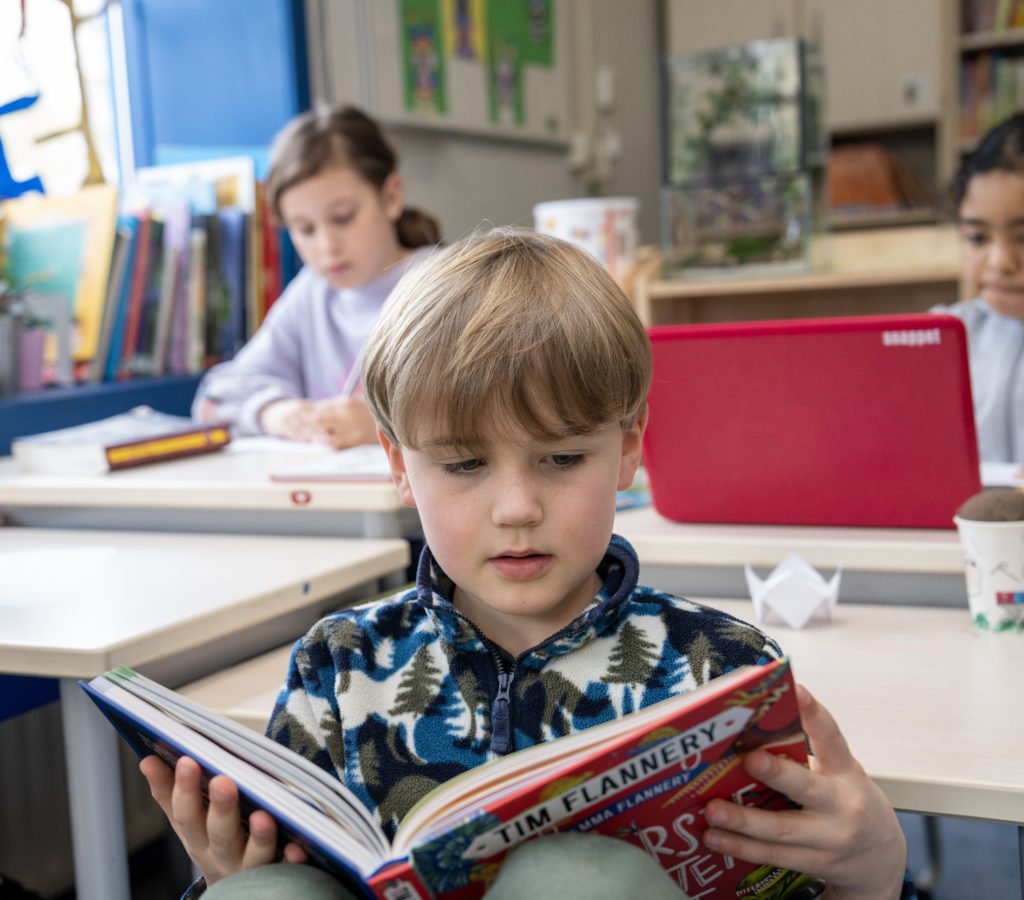 Kind leest boek, op achtergrond de rest van het klaslokaal.