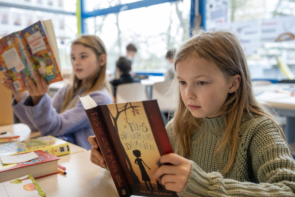 Twee meisjes die in de klas lezen in een leesboek. Een meisje heeft een paarse trui aan. Het andere meisje een kakigroene. Op de achtergrond zijn twee jongens met een activiteit bezig. 