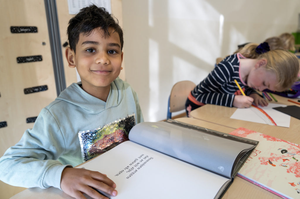 Jongen die kijkt in de camera met voor zich een boek. Naast hen zit een klasgenoot een regenboog te tekenen. 