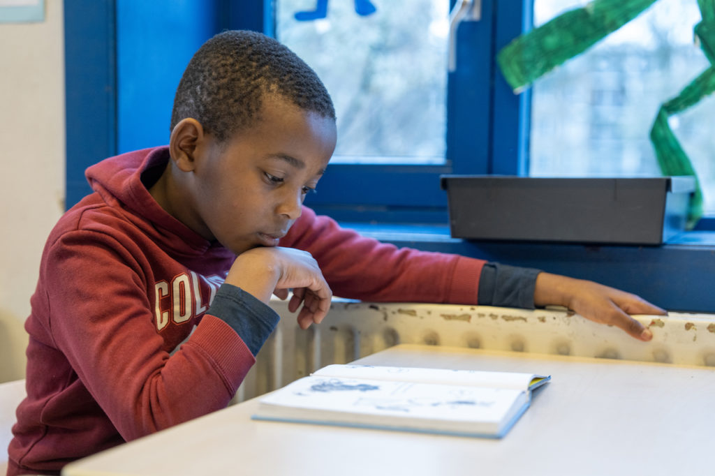 Jongen met een rode trui met donkerblauwe mouwen leest in een leesboek aan een tafel waarbij een arm op de verwarming rust. 