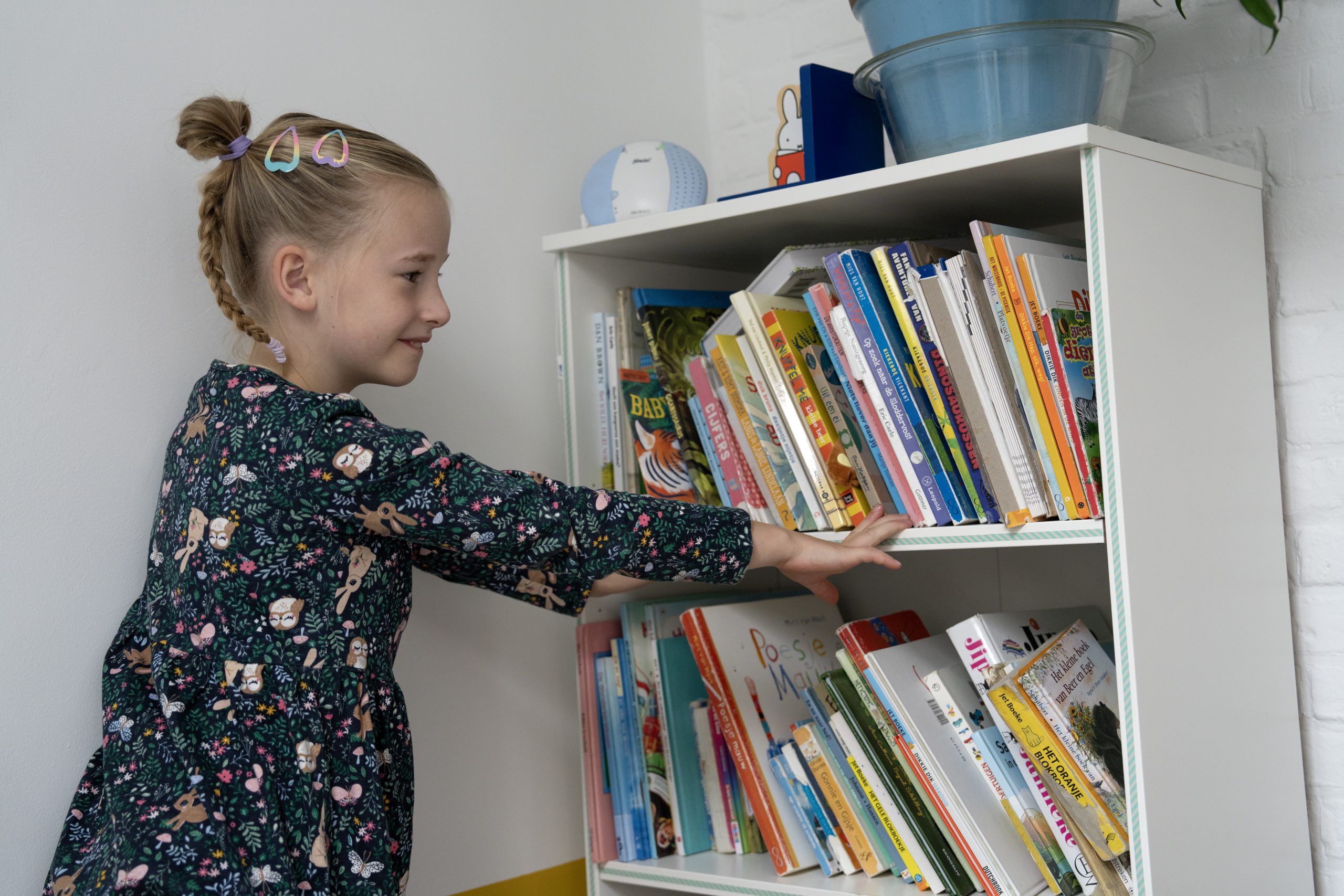 Meisje in jurkje voor een boekenkast met twee plankjes.