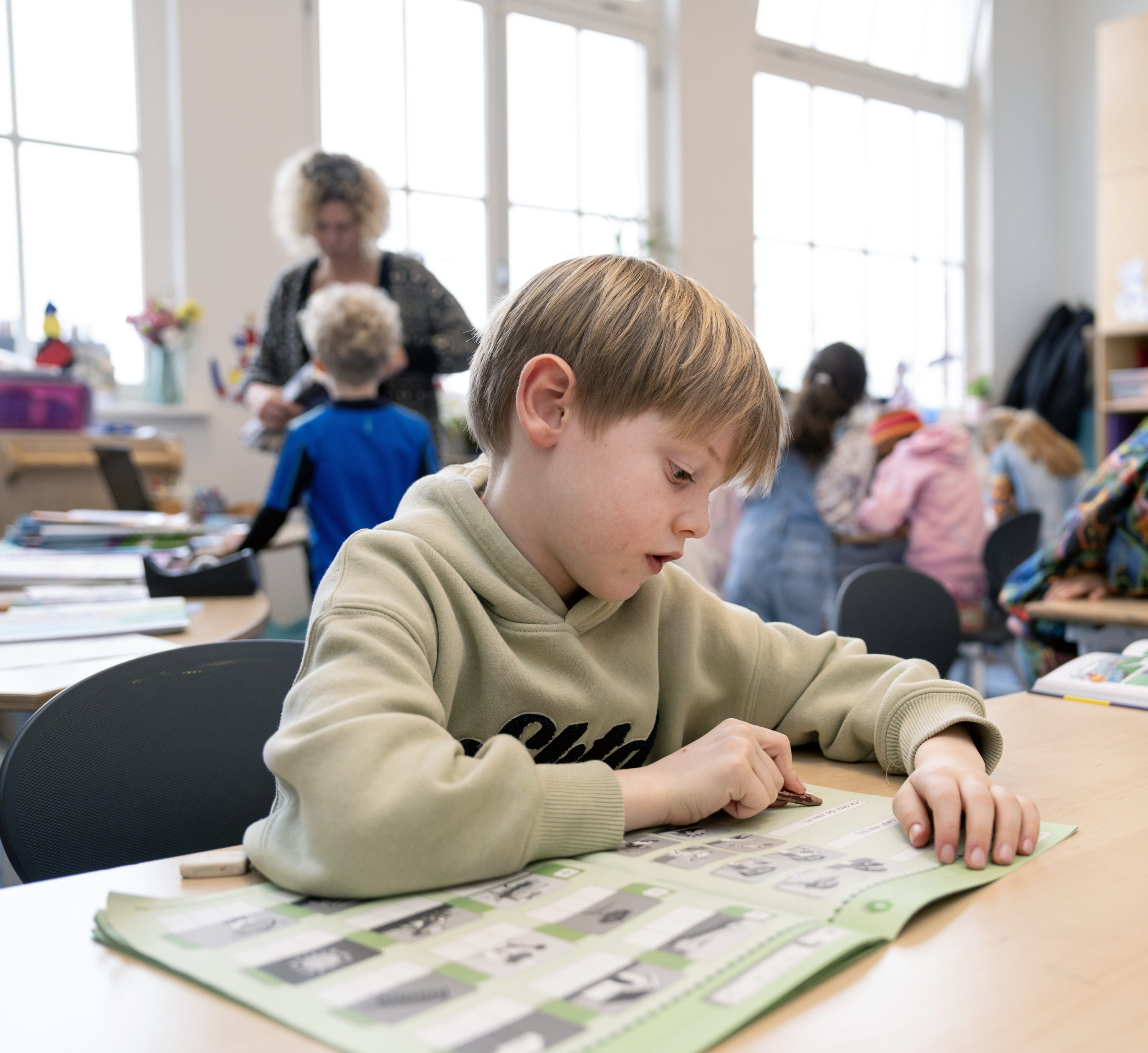 Jongetje met kakigroene trui die in de klas werkt in een werkboek van een aanvankelijk leesmethode. 